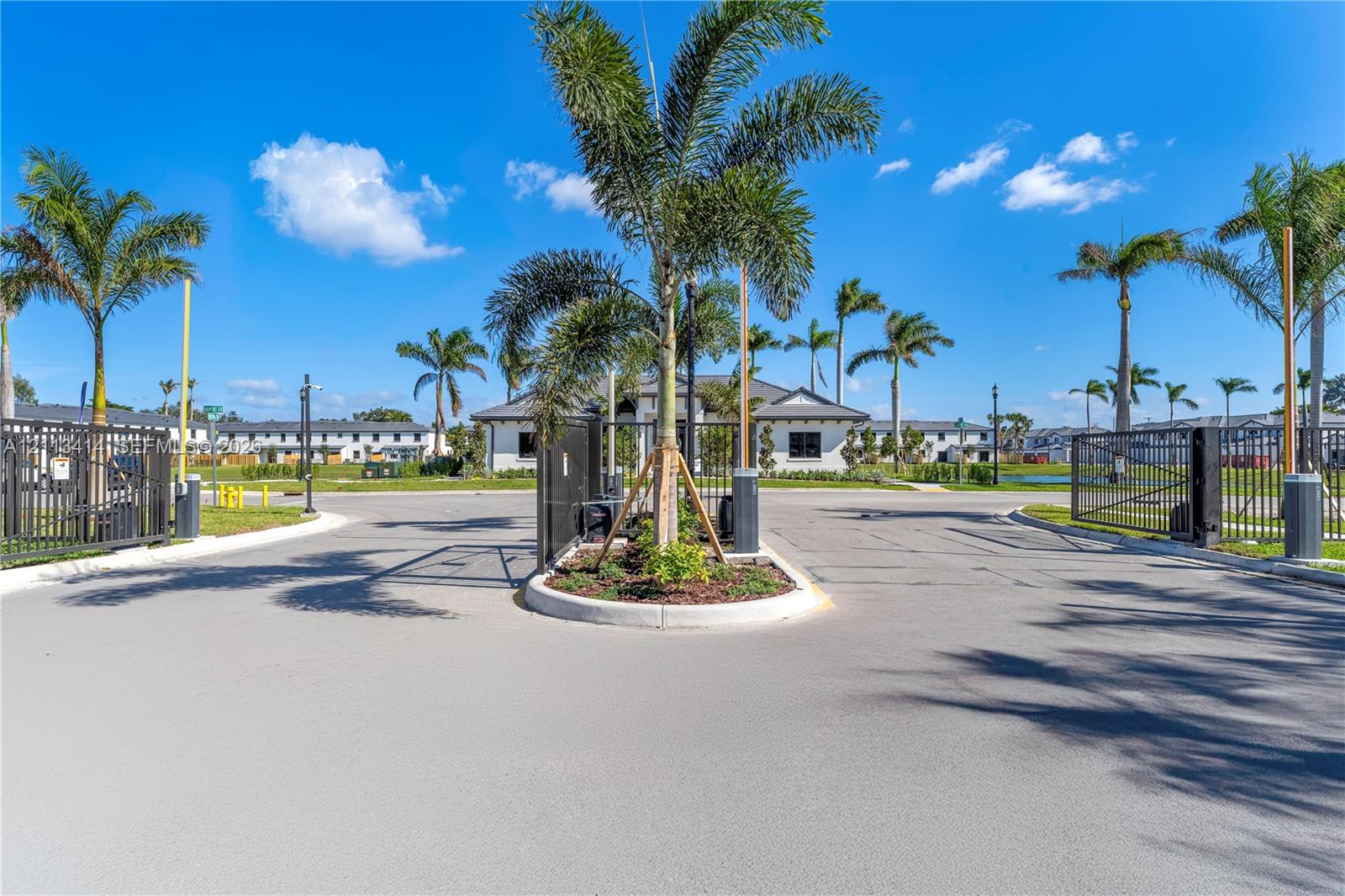 Gated community entrance featuring automated security gates, perimeter metal fencing, and palm-lined landscaping