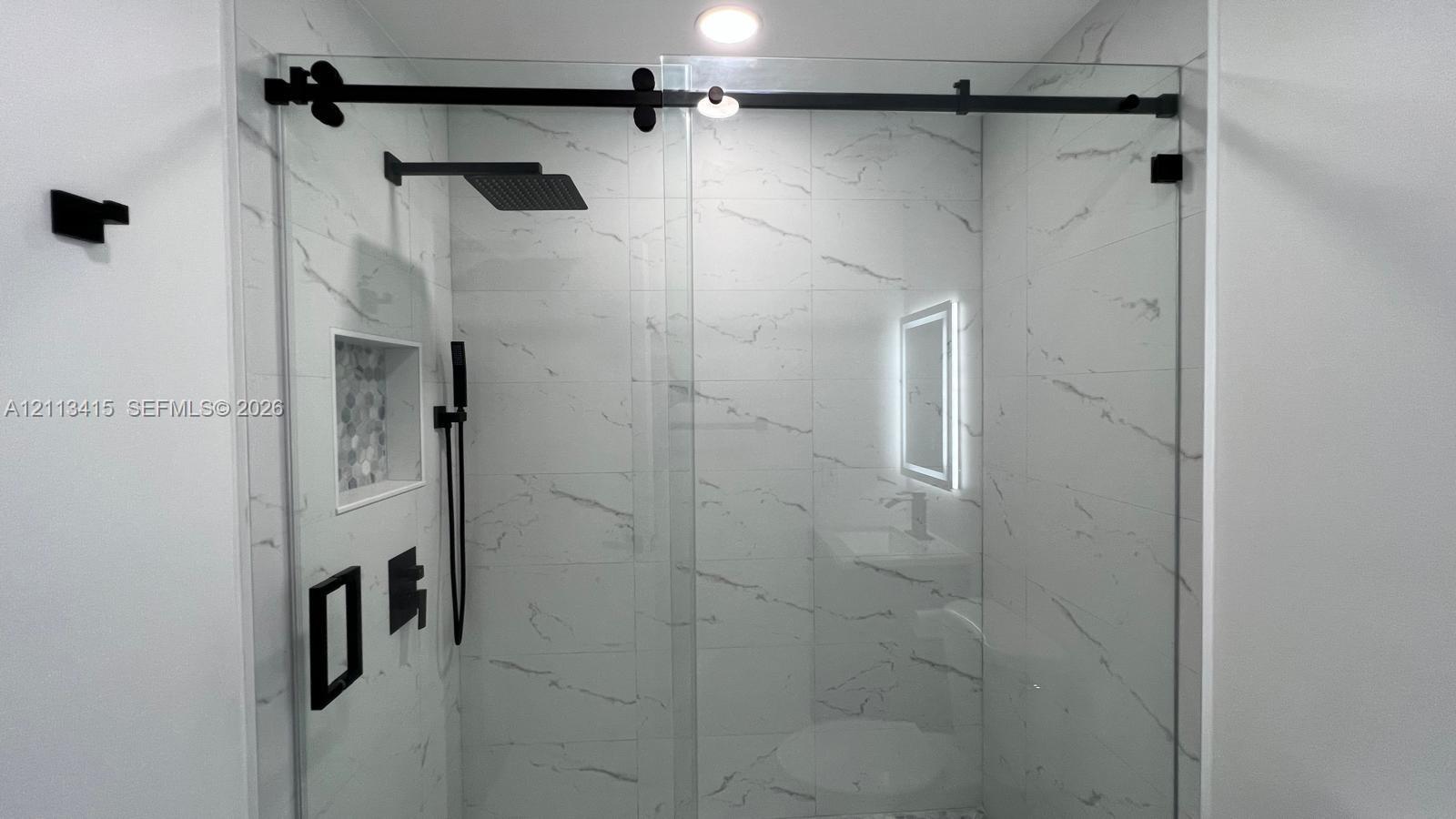 Walk-in shower featuring large-format stone-pattern tile, a glass sliding door with matte black hardware, a rainfall showerhead, and a built-in recessed storage niche