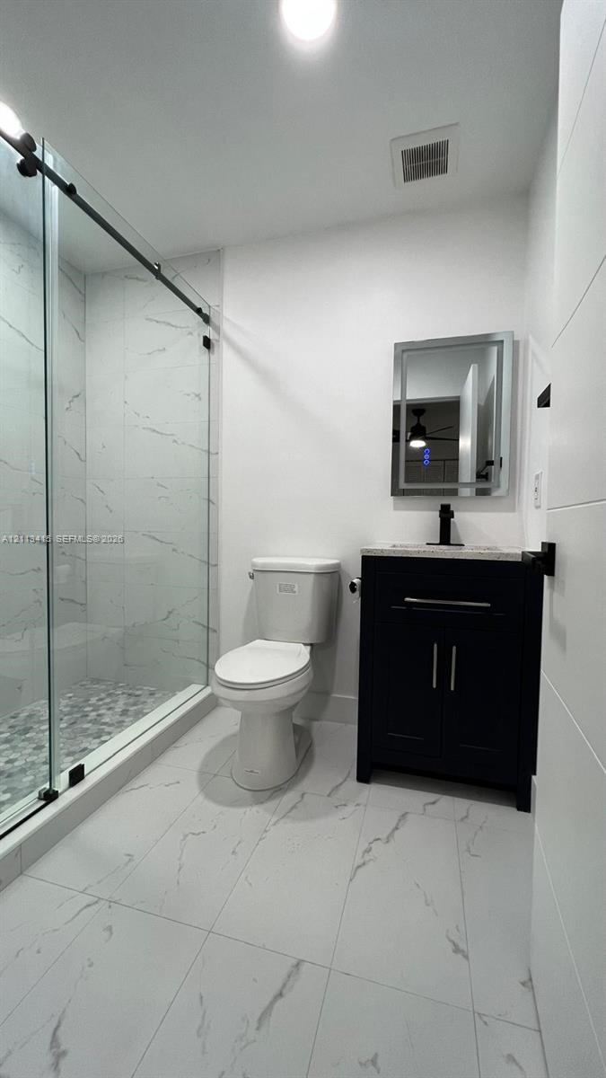 Bathroom featuring large-format tile flooring, a glass-enclosed shower with floor-to-ceiling tile surround and pebble-texture shower floor, dark cabinetry with a stone countertop, and a rectangular-framed vanity mirror
