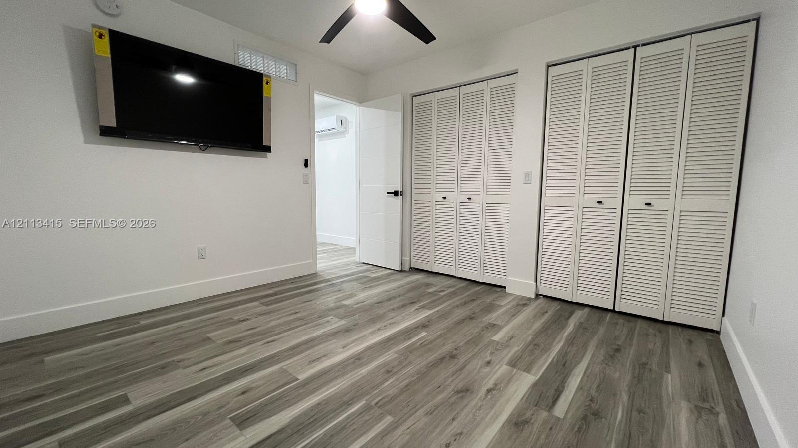 Wood-finish flooring, dual bi-fold louvered closets, matte black ceiling fan, and a wall-mounted ductless air conditioning unit