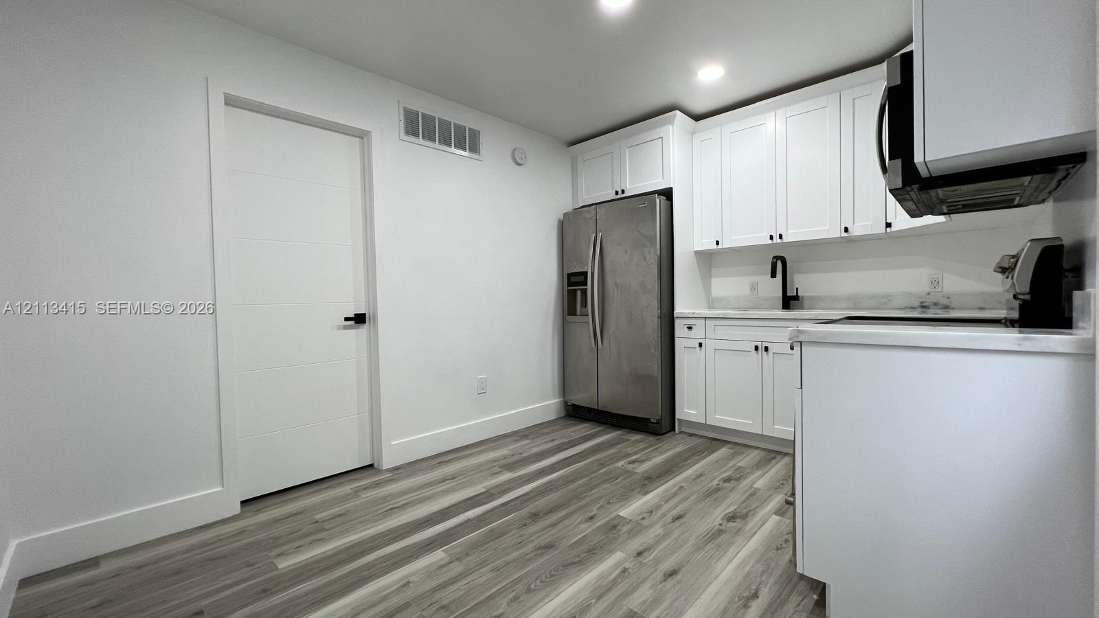 Kitchen featuring white Shaker-style cabinetry, light-toned stone-finish countertops, matte black plumbing fixtures, stainless steel refrigerator, and wood-finish flooring