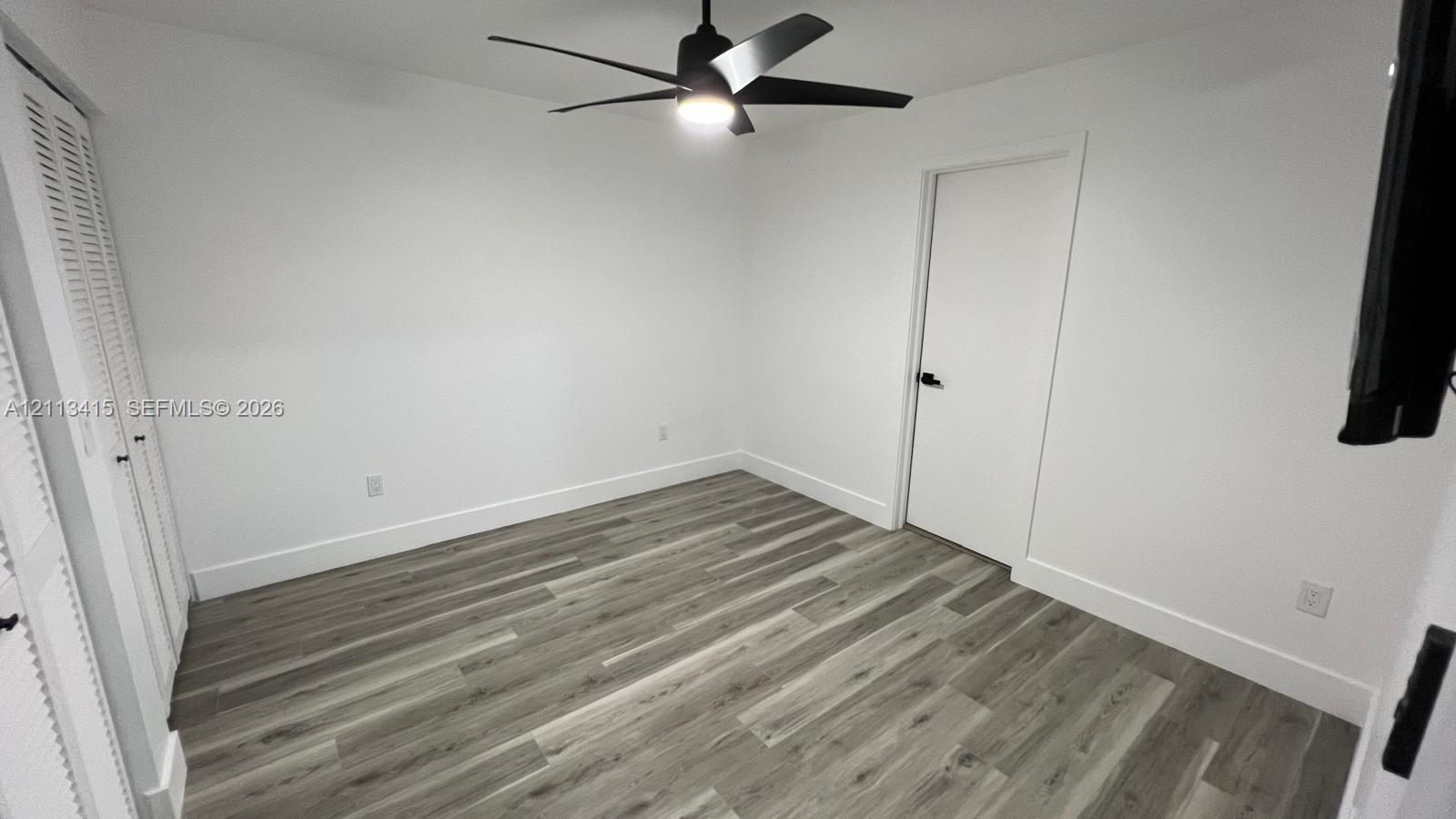 Room featuring wood-finish flooring, white walls, dual louvered-panel closet doors, and a matte black ceiling fan with an integrated light fixture