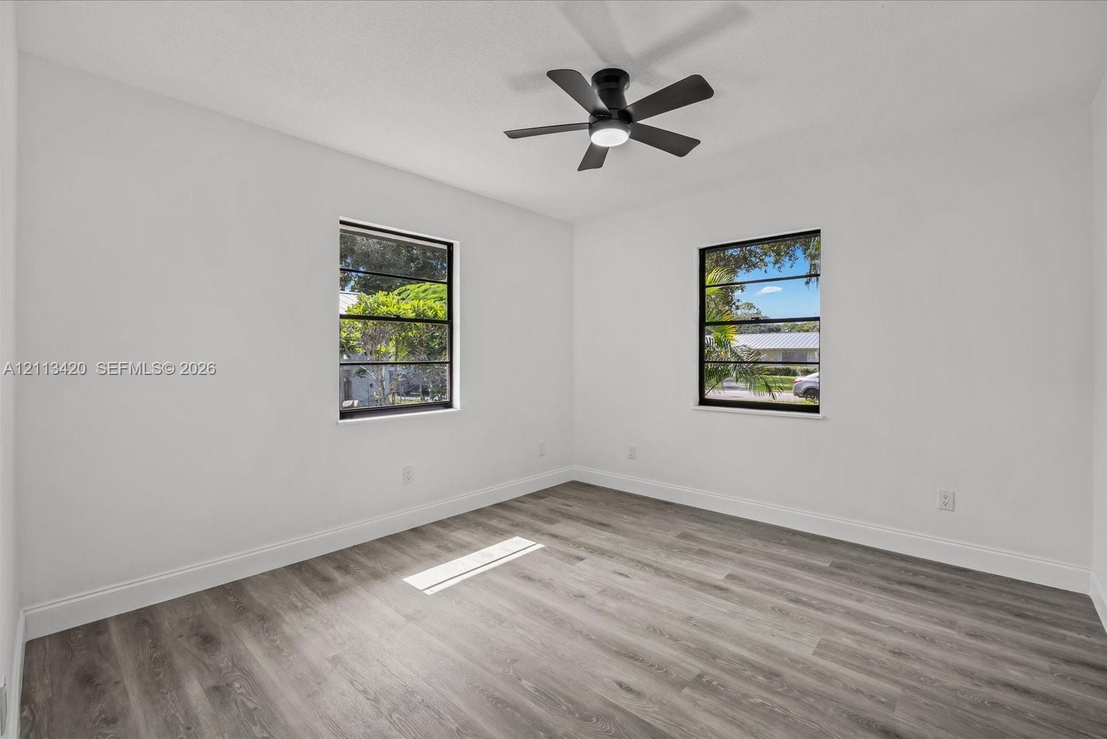 Neutral-toned interior room featuring wood-finish flooring, two windows with black frames, and a ceiling fan with integrated lighting