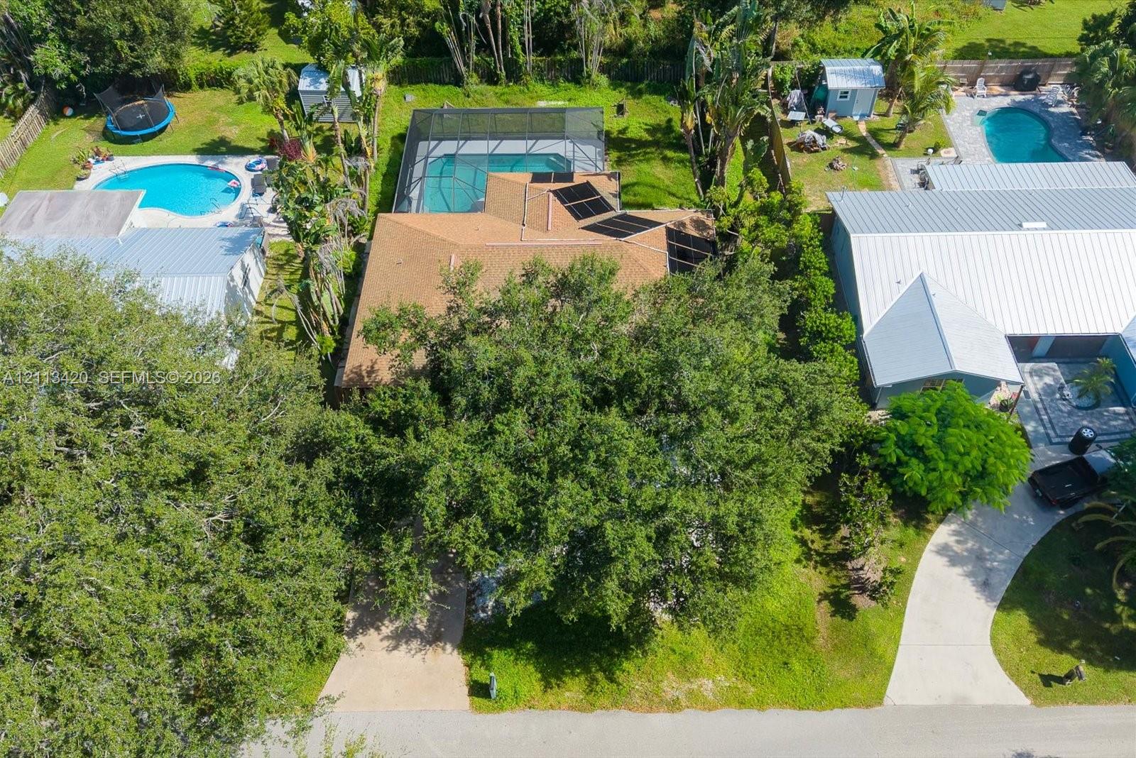Aerial perspective of a property featuring an asphalt shingle roof with integrated solar panels, a screened-in swimming pool enclosure, mature canopy trees, and a paved concrete driveway