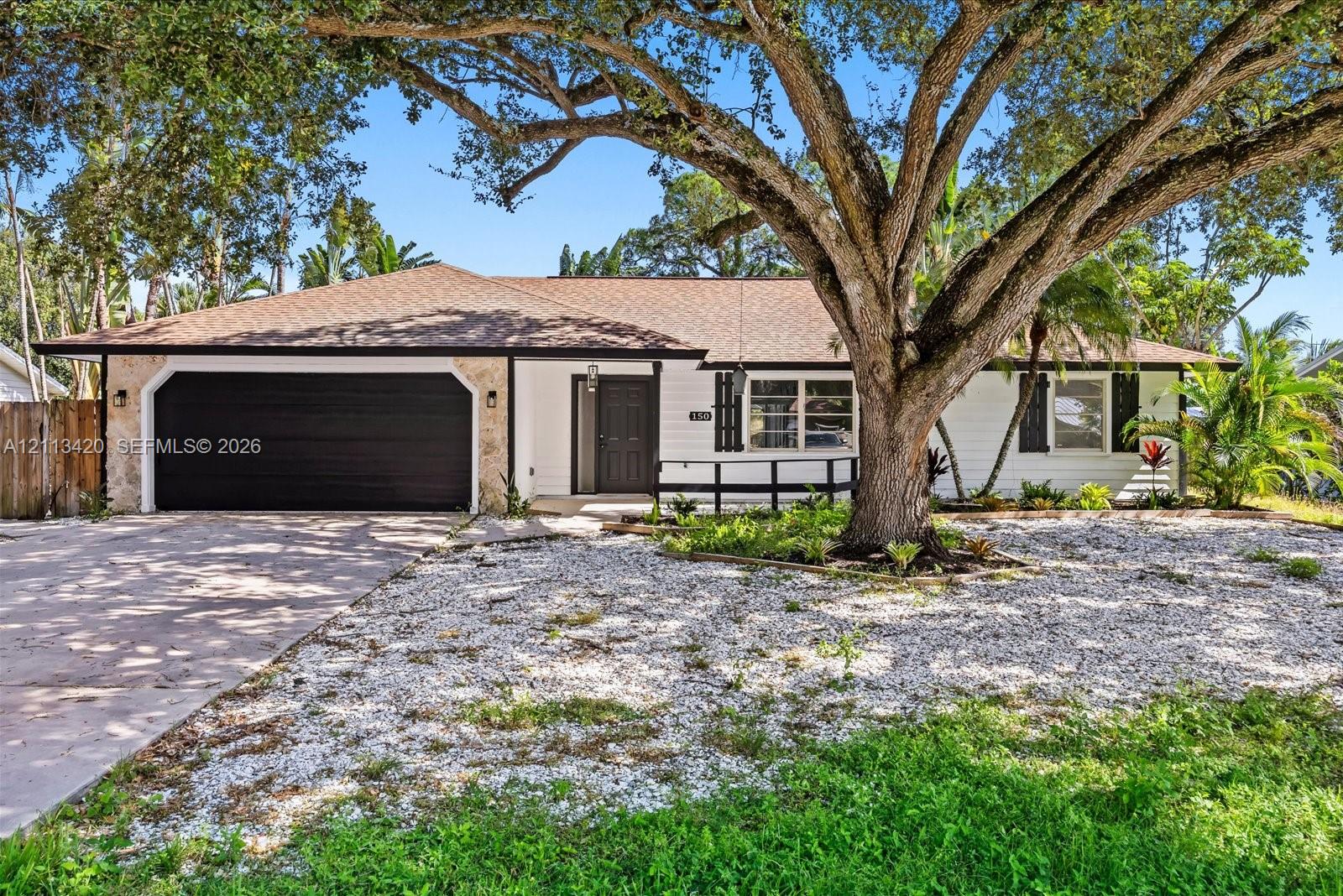 Single-story exterior featuring white horizontal siding, stone-accented garage facade, dark-toned double-car garage door, and mature canopy tree