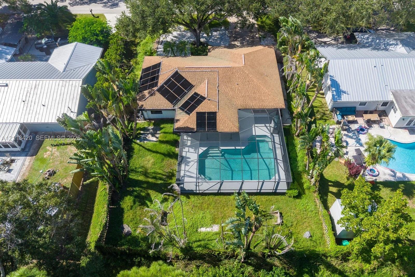 Aerial perspective of a property featuring an in-ground pool enclosed within a screened lanai, roof-mounted solar panels, mature landscaping, and a fenced perimeter