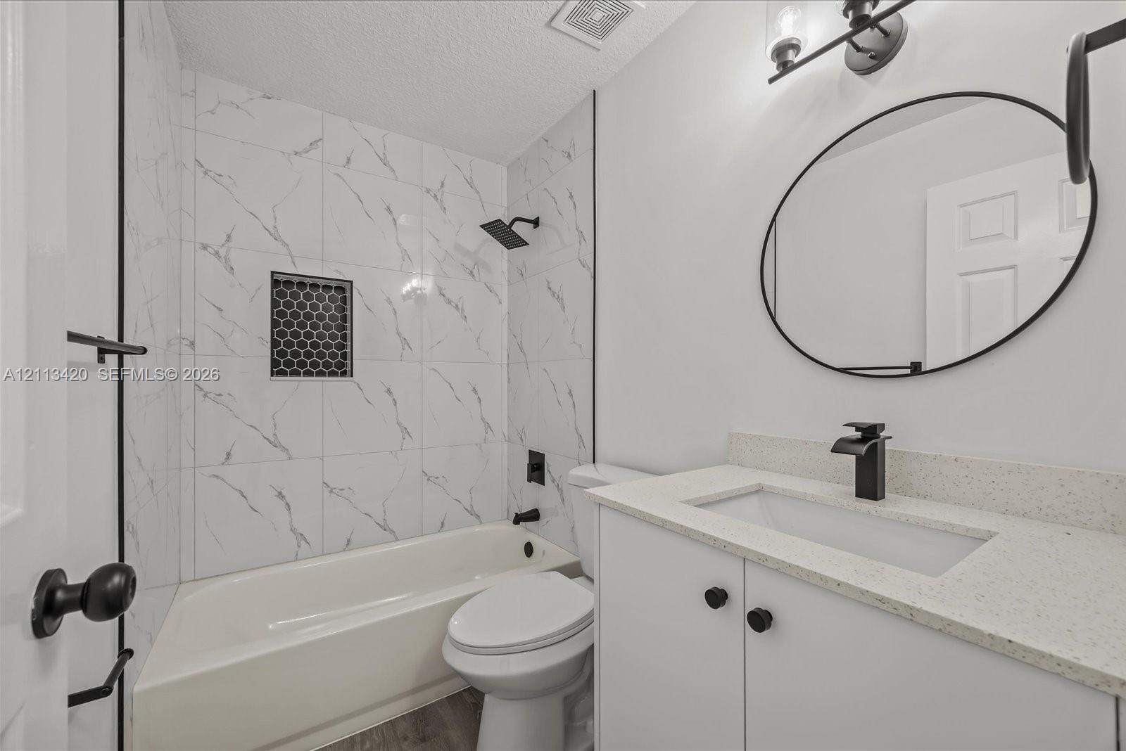 Bathroom featuring stone-finish wall tile in the tub surround with a hexagon-tile niche, matte black rainfall shower fixture, quartz-surround vanity, oval wall-mounted mirror, and matte black faucet hardware