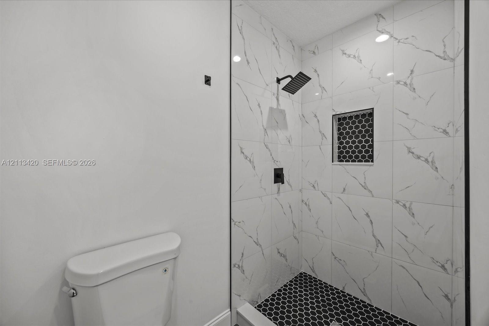 Walk-in shower featuring large-format veined wall tile, matte black rainfall shower head, recessed hexagonal tile niche, and contrasting hexagonal floor tile