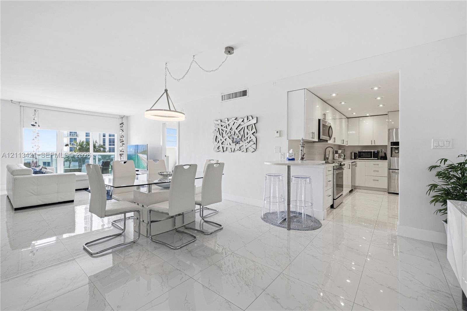 Open-concept area featuring white high-gloss cabinetry, stainless steel appliances, polished stone-finish floor tiling, a peninsula with a breakfast bar, and a modern circular pendant light fixture