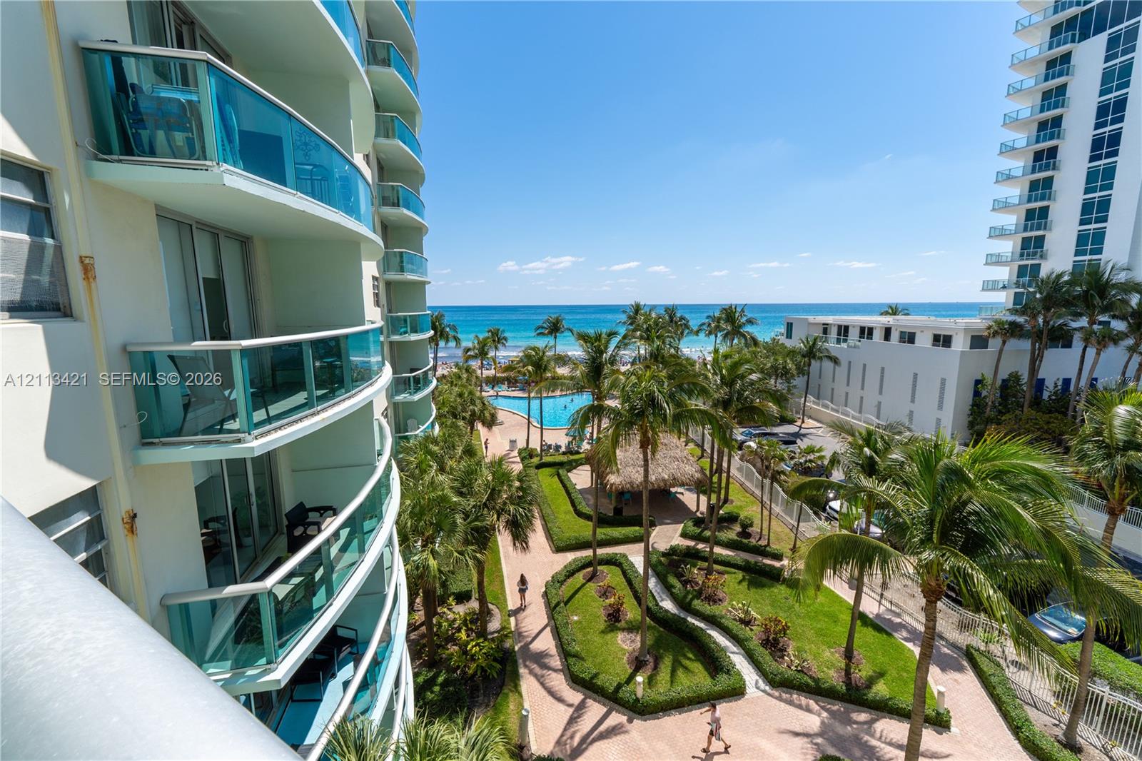 High-rise building with glass-paneled curved balconies, landscaped courtyards with paved pathways, an outdoor swimming pool, and ocean scenery
