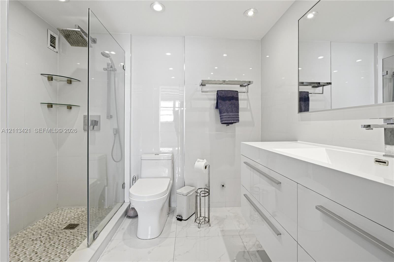 Modern bathroom featuring a glass-enclosed walk-in shower with rainfall showerhead, pebble-tiled shower floor, gloss-finish floating vanity with integrated sink, and recessed lighting