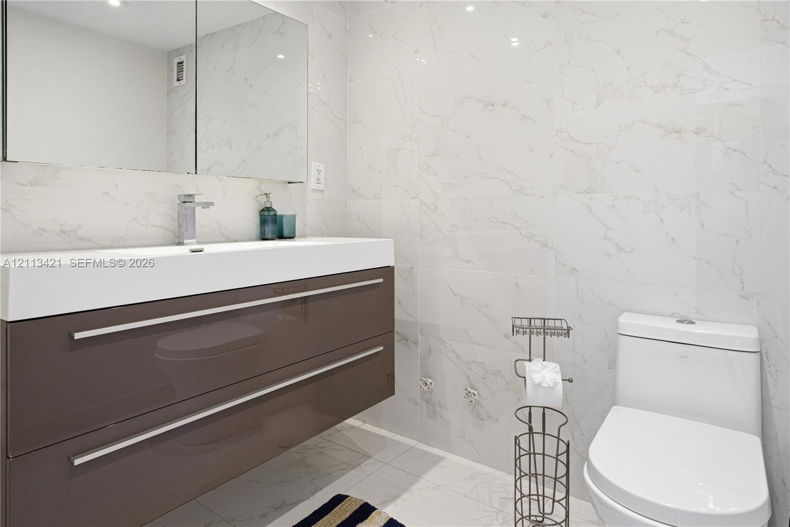 Floating vanity featuring a high-gloss finish, integrated sink, and polished chrome hardware, paired with floor-to-ceiling stone-patterned tile walls, a mirrored medicine cabinet, and a contemporary one-piece toilet Floating vanity featuring a high-gloss finish, integrated sink, and polished chrome hardware, paired with floor-to-ceiling stone-patterned tile walls, a mirrored medicine cabinet, and a contemporary one-piece toilet