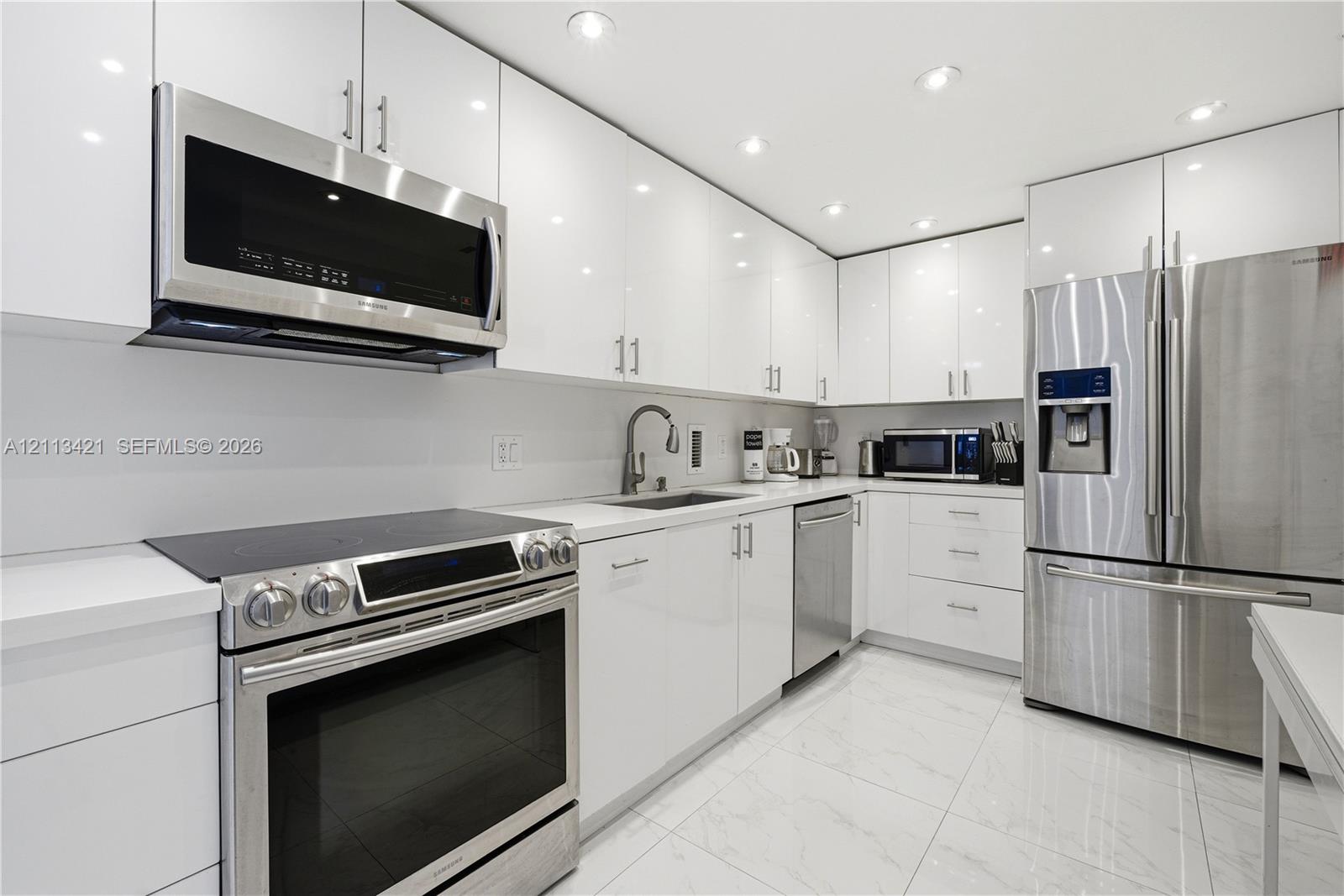 Modern kitchen featuring high-gloss white cabinetry, quartz countertops, stainless steel appliances, and large-format tile flooring