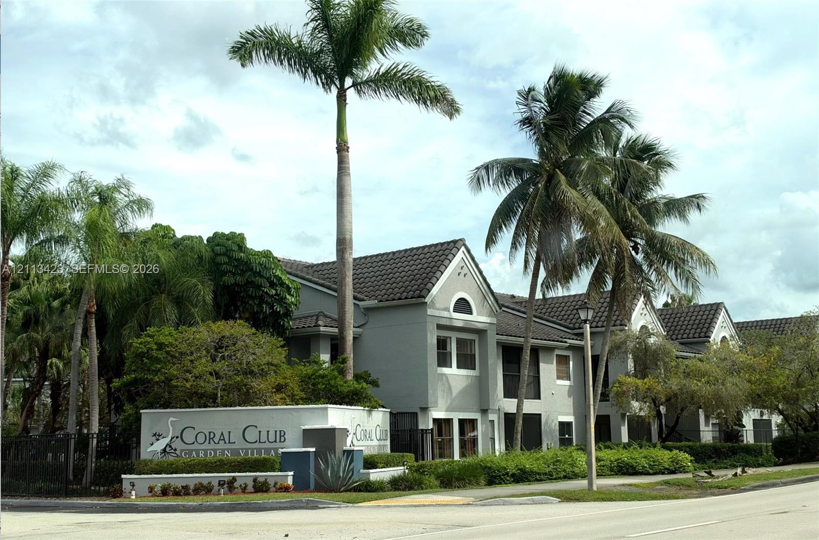 Multi-unit stucco building featuring a tile roof, screened-in balconies, mature palm trees, and professionally manicured landscaping