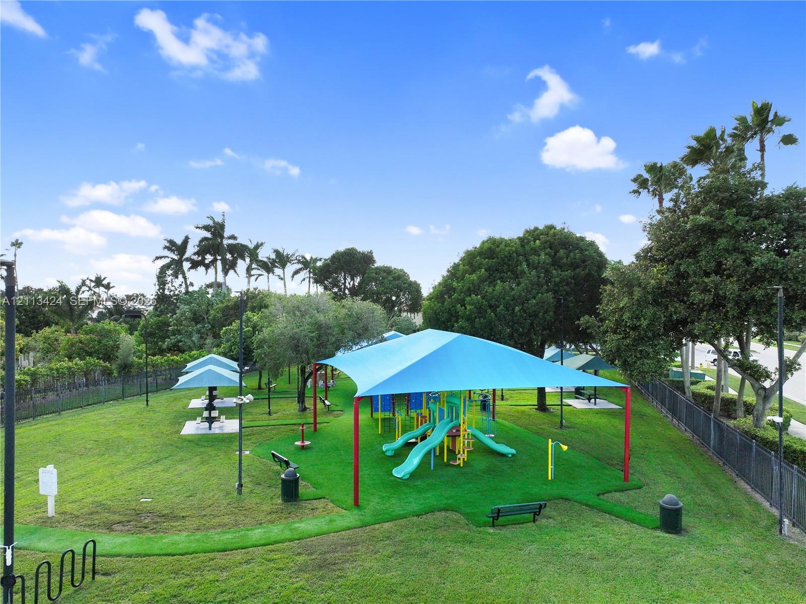 Outdoor recreation area featuring a playground structure with a blue shade canopy, integrated turf surfacing, picnic stations, perimeter fencing, and mature landscaping