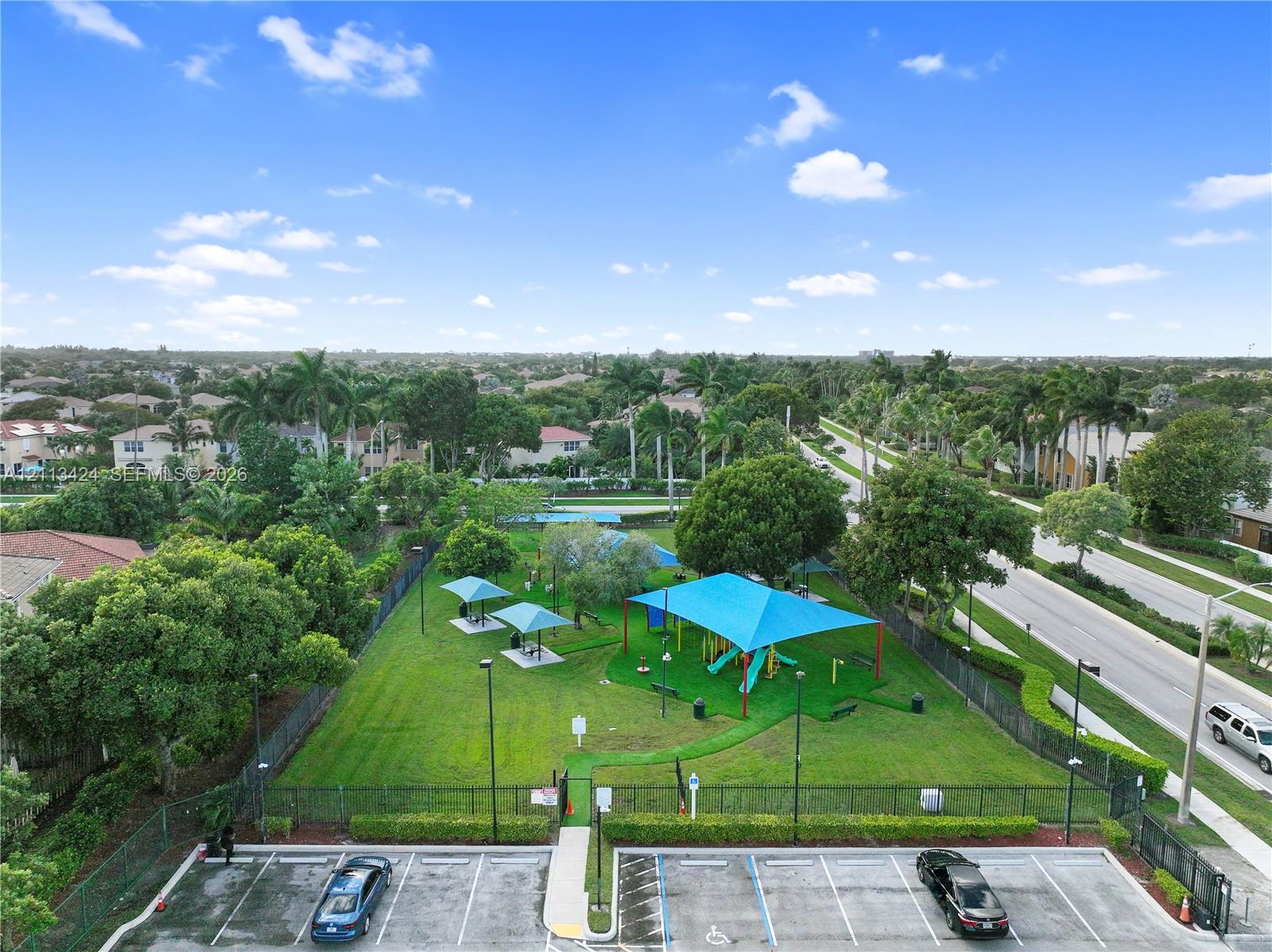 Community park area featuring a fenced perimeter, expansive lawn, multiple shaded structures, and designated paved parking