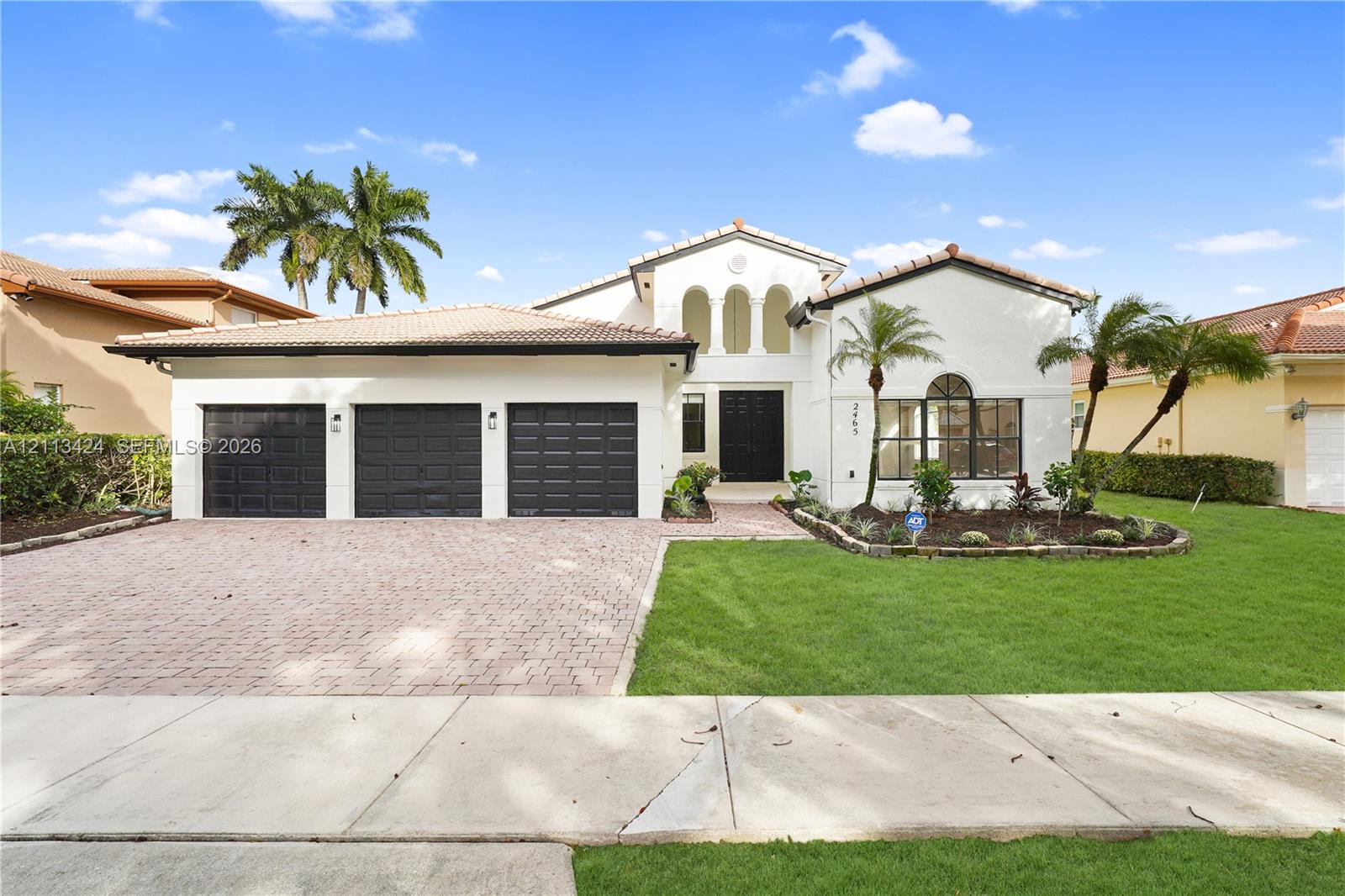 Stucco exterior featuring a tile roof, three-car garage with paneled doors, arched entryway, and a brick-paver driveway (virtually edited out tree)