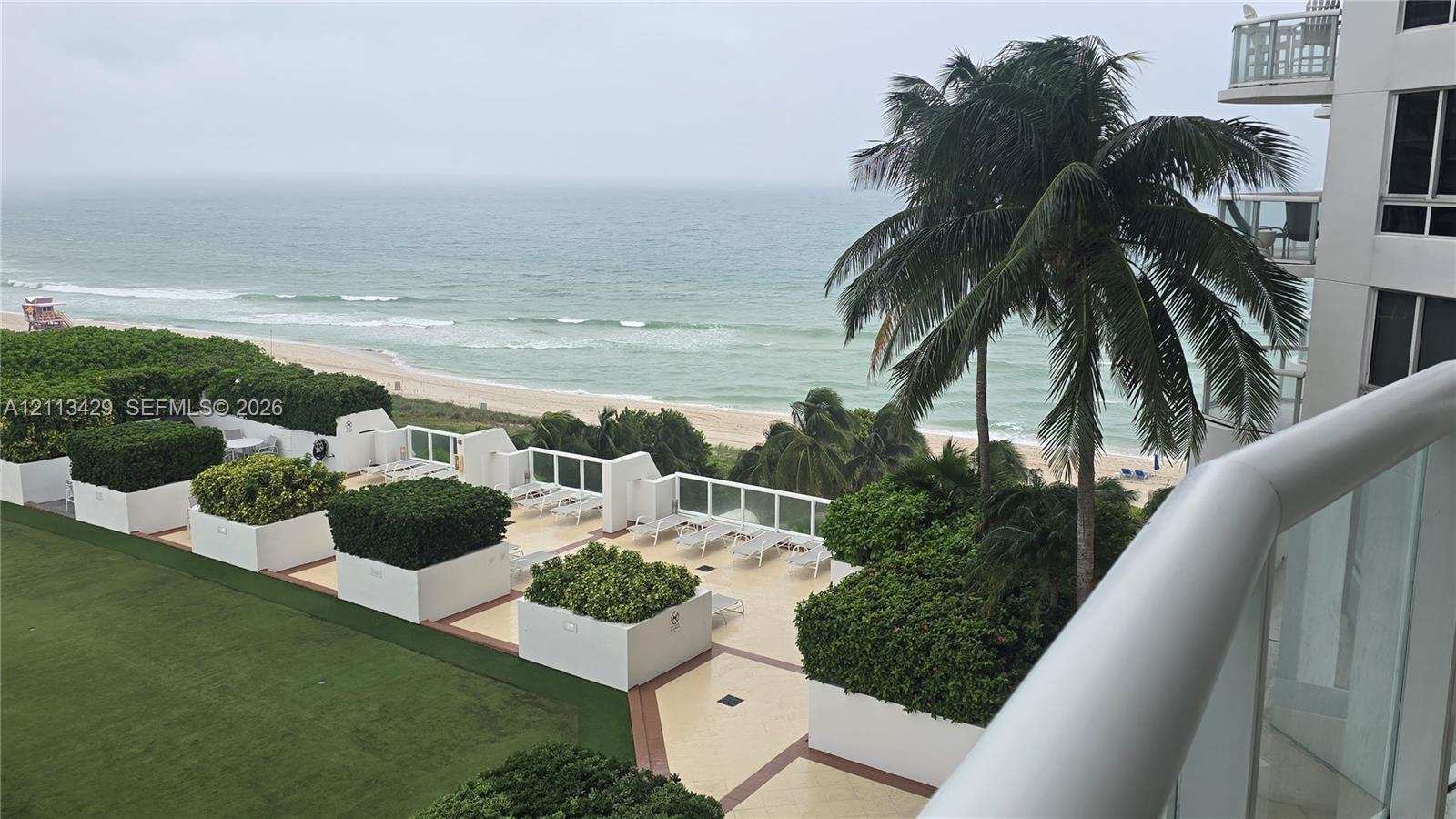 Elevated terrace featuring white glass-panel railings, tile flooring, built-in planter boxes with mature greenery, and direct oceanfront scenery