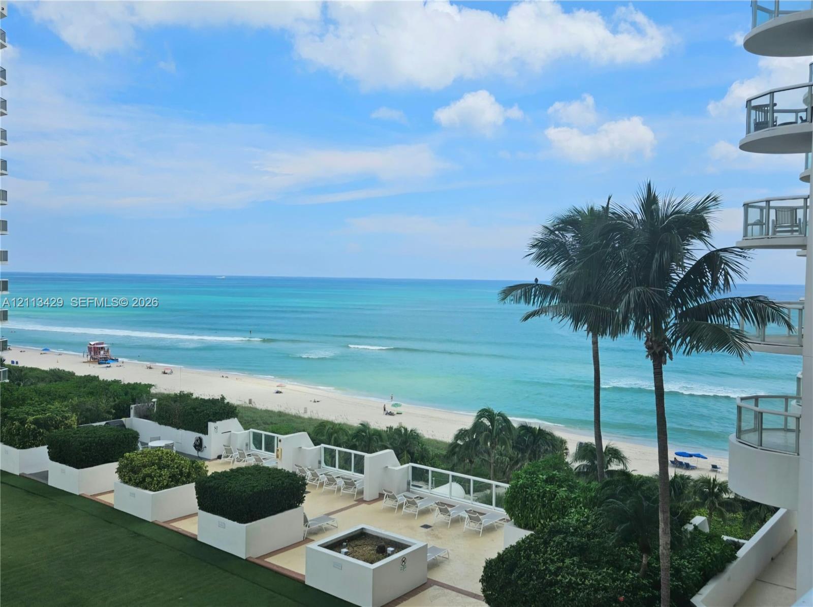 Elevated terrace featuring a stone-tile floor, built-in concrete planters with mature greenery, and metal-railing balconies overlooking the ocean and sandy coastline Elevated terrace featuring a stone-tile floor, built-in concrete planters with mature greenery, and metal-railing balconies overlooking the ocean and sandy coastline