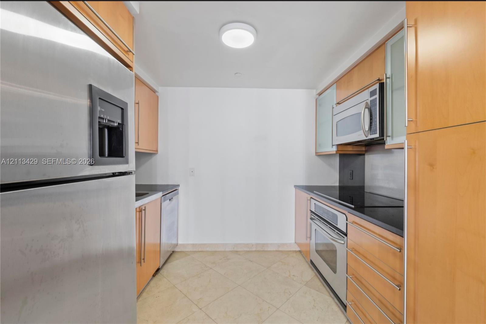 Galley kitchen featuring light wood cabinetry, stone countertops, stainless steel appliances, and tile flooring