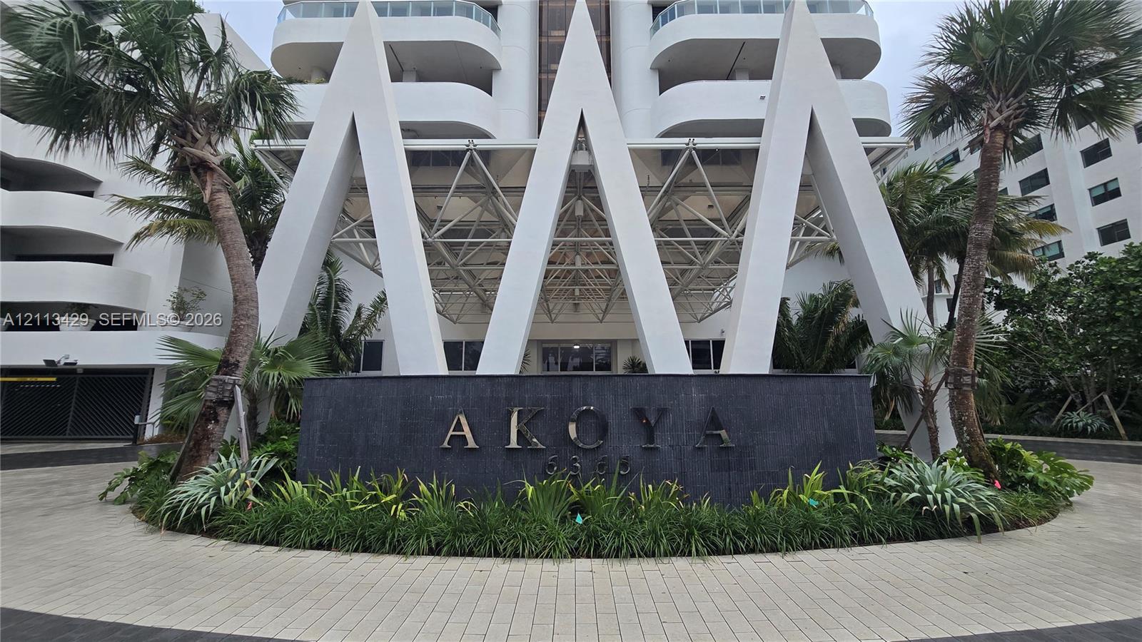 High-rise exterior featuring prominent white geometric structural detailing, a dark stone entrance monument with architectural signage, a paved circular motor court, and mature palm landscaping