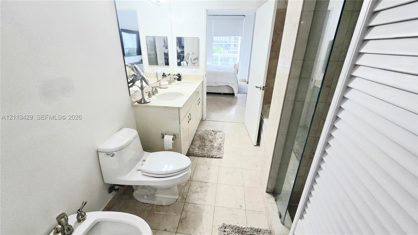Bathroom featuring a dual-sink vanity with light-toned cabinetry, stone countertop, wall-to-wall mirror, glass-enclosed shower, and tile flooring