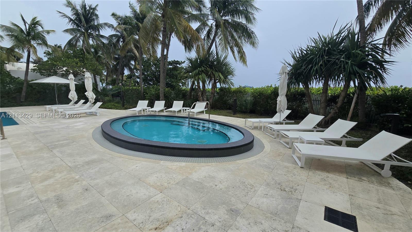 Oval-shaped spa featuring a dark tiled perimeter, surrounded by light-toned natural stone pavers, integrated pool edge, and mature palm trees
