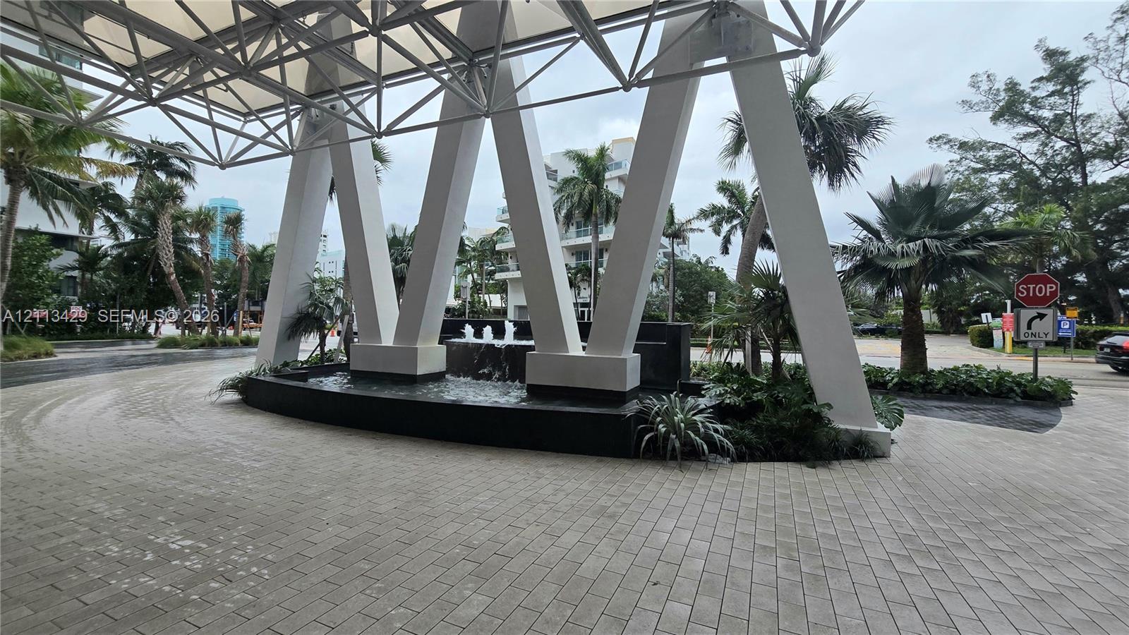 Covered porte-cochre featuring geometric structural supports, stone-paver driveway, and a built-in water feature with lush landscaping