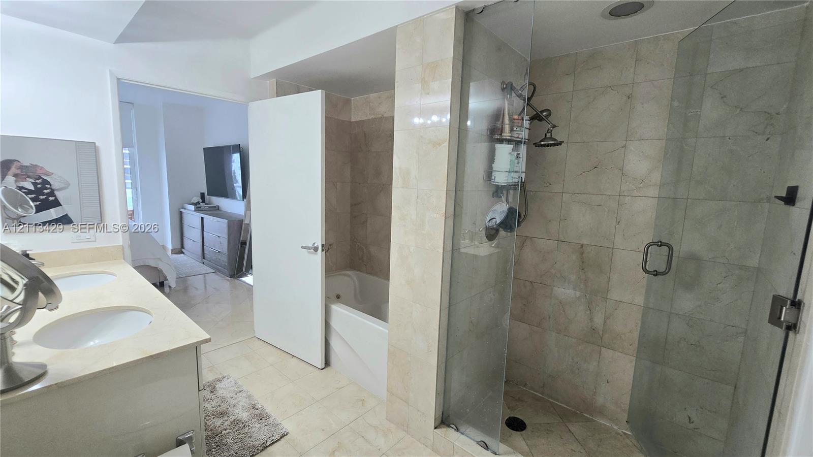 Dual-sink vanity with light-toned countertop, soaking tub, glass-enclosed shower with stone-tile surround, and tiled flooring
