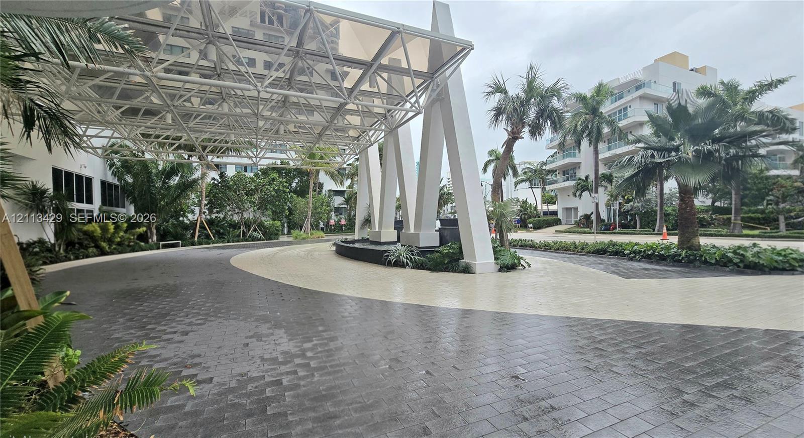Covered porte-cochre featuring a structural geometric canopy, decorative stone masonry fountain, paved circular driveway, and manicured tropical landscaping