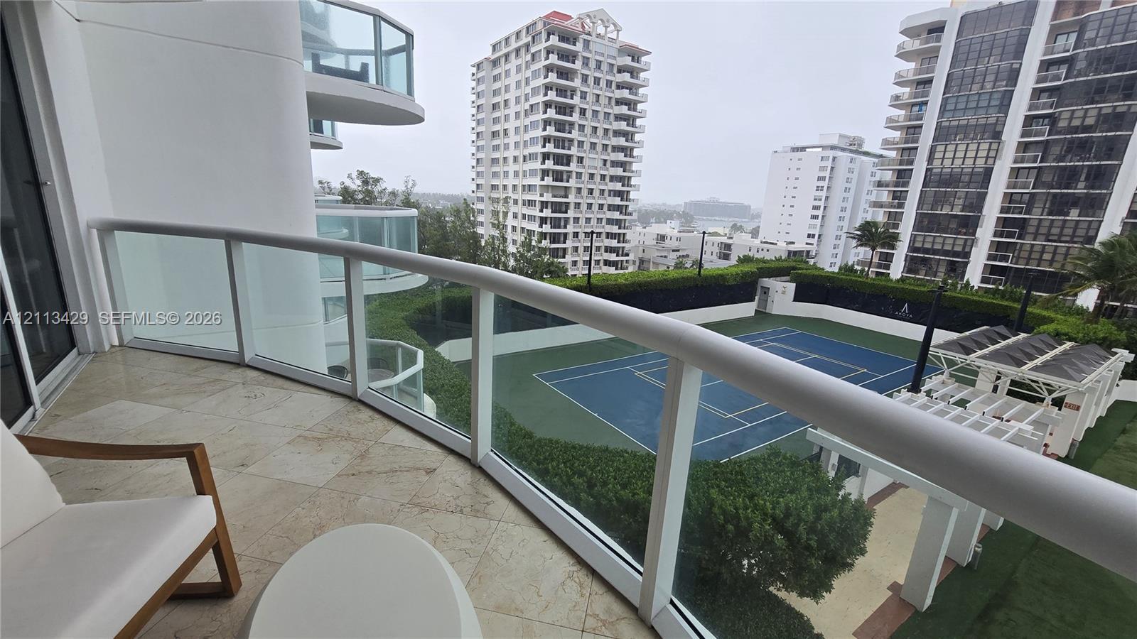 Covered balcony featuring stone-tile flooring and a curved glass railing overlooking a tennis court