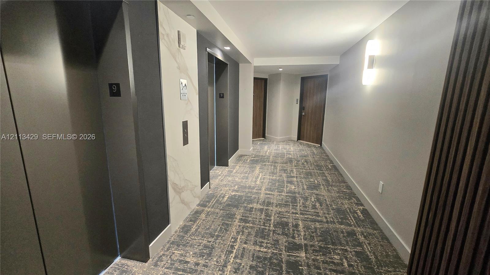 Hallway featuring patterned wall-to-wall carpeting, stone-look elevator surround, recessed lighting, and wall-mounted sconces