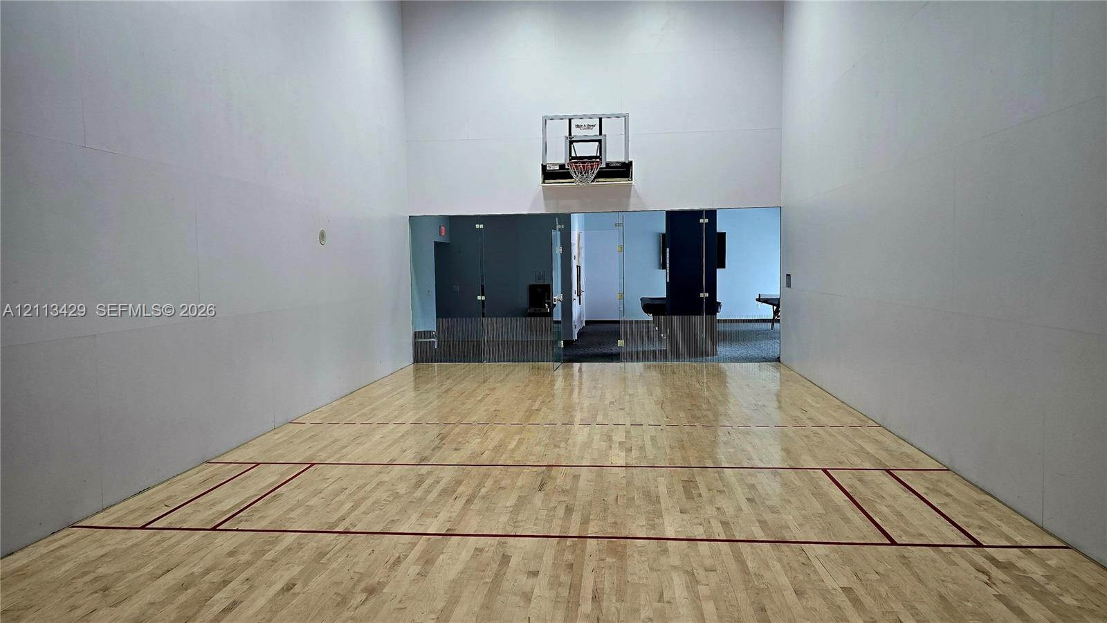 Indoor court featuring wood-finish flooring, high ceilings, integrated wall-mounted basketball hoop, and glass-paneled partition