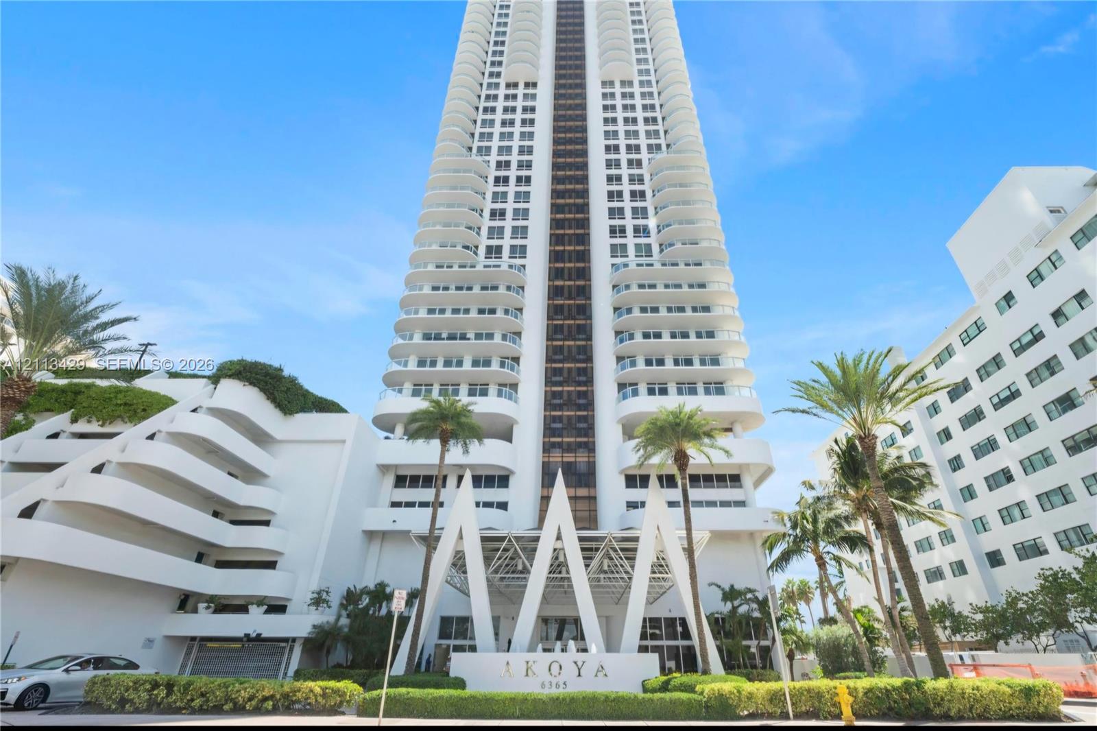 High-rise residential tower featuring a white facade, structural balconies, geometric entryway architecture, and established landscaping