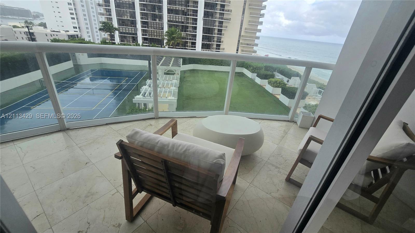 Balcony featuring stone tile flooring and a glass-paneled railing, overlooking a hard-surfaced tennis court, a manicured lawn, and a white architectural pergola