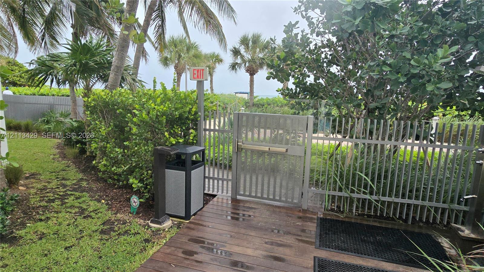 Outdoor area featuring a wood-finish deck, metal perimeter fencing with a gated entry, manicured landscaping, and mature palm trees