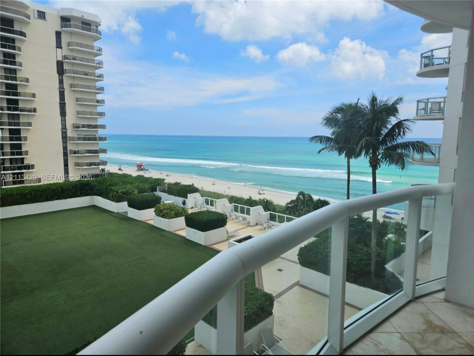 Private balcony featuring a glass panel railing, stone tile flooring, an expansive synthetic turf terrace, built-in planter boxes with integrated landscaping, and oceanfront exposure