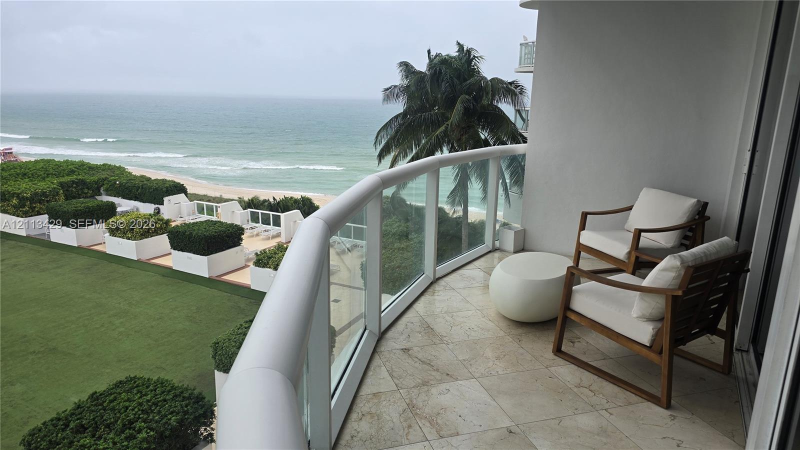 Covered balcony featuring stone tile flooring and a glass panel railing overlooking the oceanfront and manicured grounds