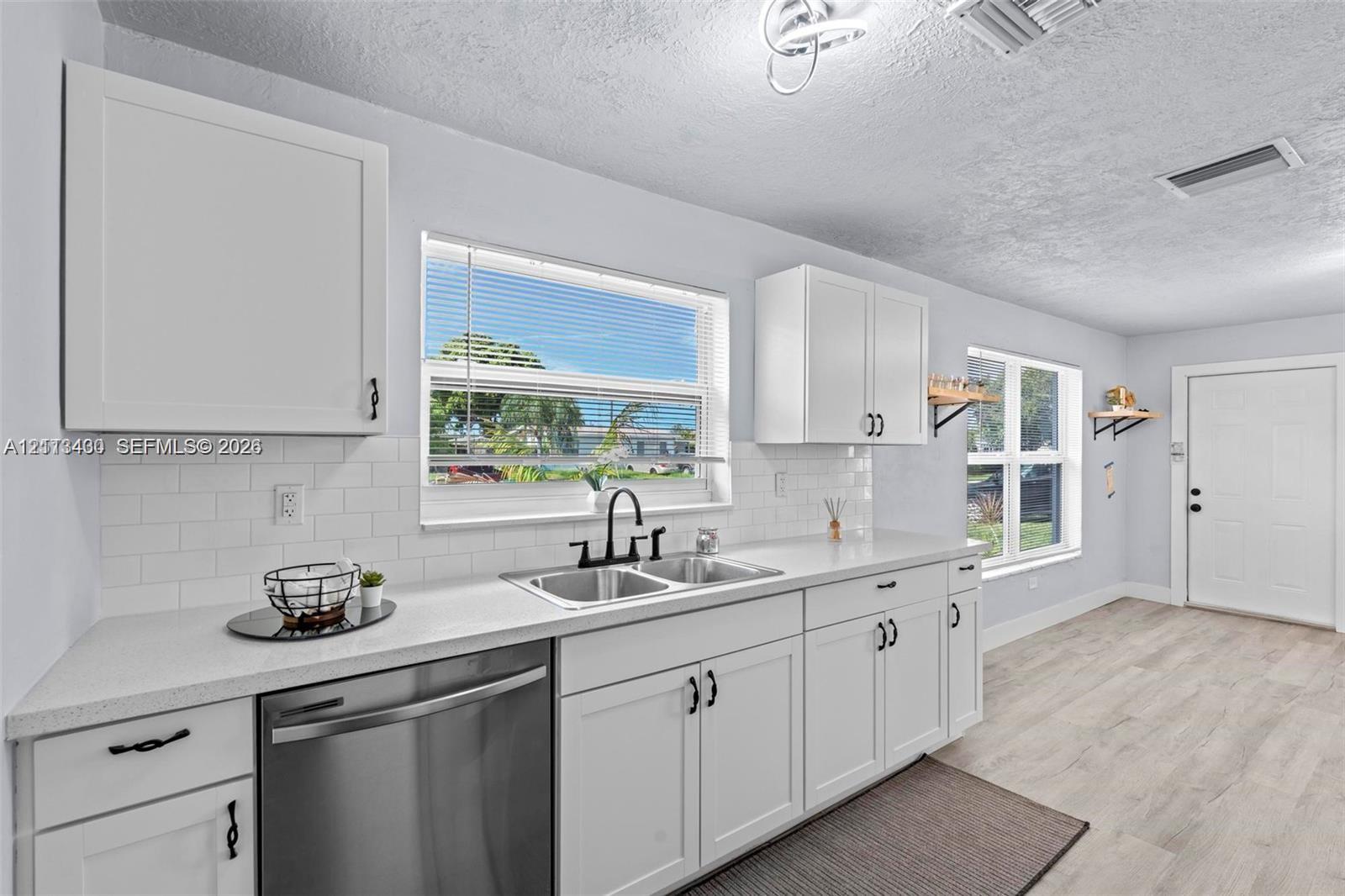 Kitchen featuring white shaker-style cabinetry, white subway tile backsplash, solid-surface countertops, dual-basin stainless steel sink with matte black hardware, and wood-finish flooring Kitchen featuring white shaker-style cabinetry, white subway tile backsplash, solid-surface countertops, dual-basin stainless steel sink with matte black hardware, and wood-finish flooring