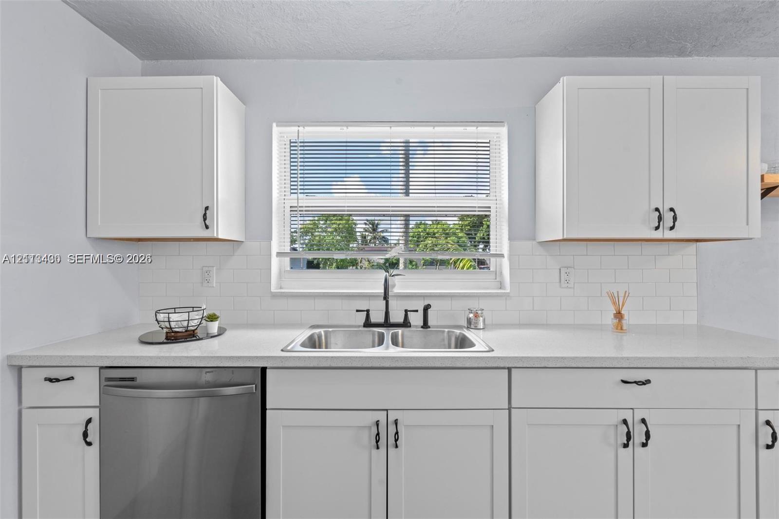 White shaker-style cabinetry, stainless steel dishwasher, white subway tile backsplash, solid-surface countertop, and dual-basin stainless steel sink with matte black hardware