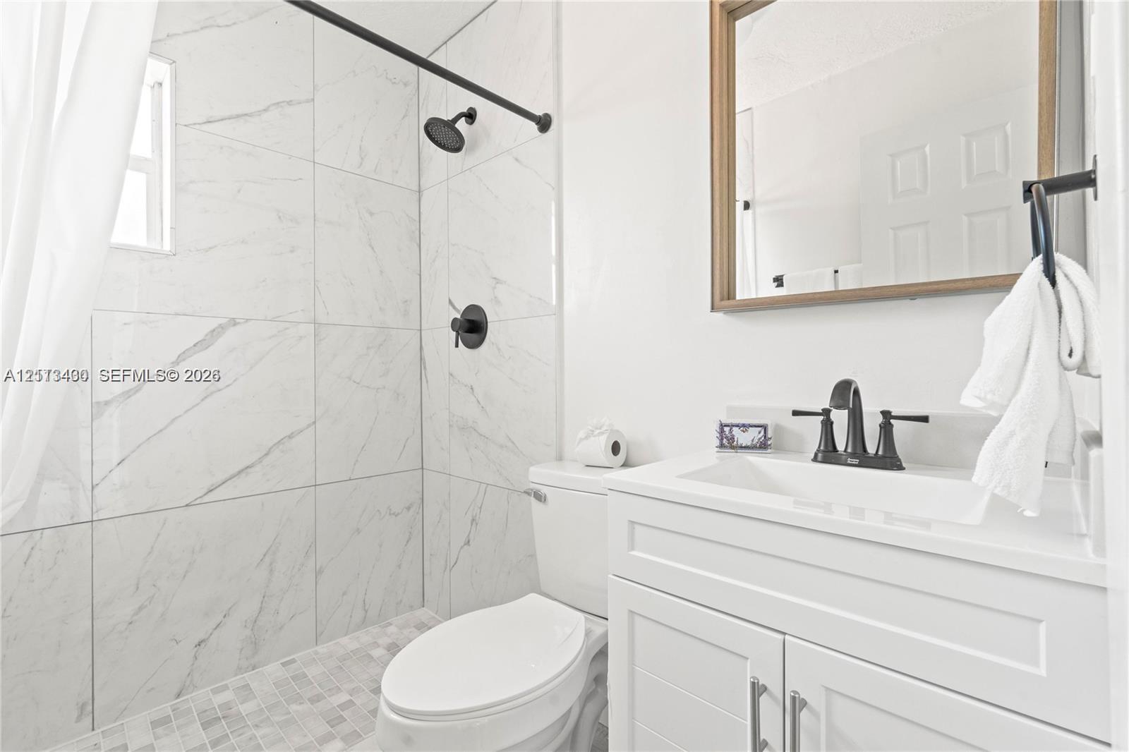 Bathroom featuring floor-to-ceiling large-format tile shower surround, matte black rainfall shower head, white vanity cabinet with shaker-style doors, and rectangular undermount sink with widespread matte black faucet