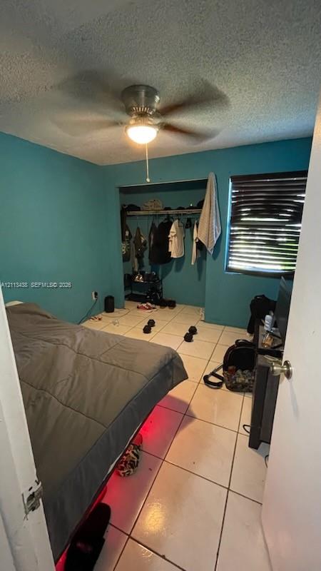 Room featuring white ceramic tile flooring, a built-in closet with overhead shelving, a ceiling fan with integrated lighting, and horizontal window blinds