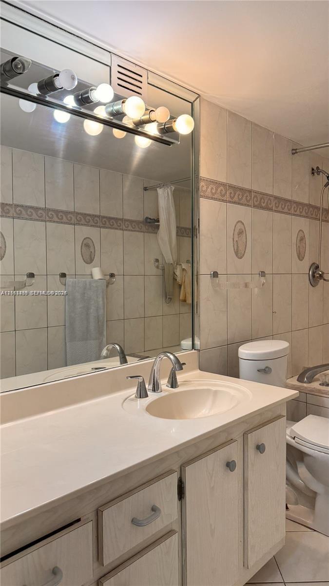 Bathroom featuring a vanity with integrated sink, expansive wall-to-wall mirror, multi-bulb overhead lighting, and full-height tiled walls with decorative trim