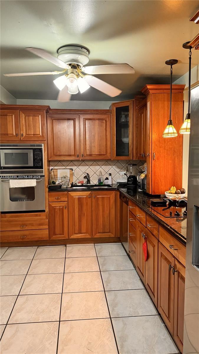 Kitchen featuring medium-tone wood cabinetry, dark stone countertops, stainless steel wall oven and microwave, ceramic tile flooring, and dual glass pendant light fixtures