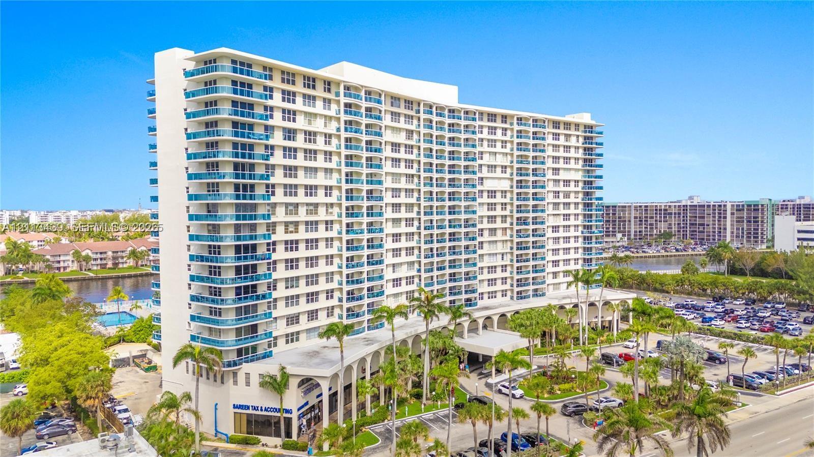 High-rise residential building featuring a curved architectural design, stacked glass-railed balconies, a ground-level colonnade, and landscaped grounds with palm trees