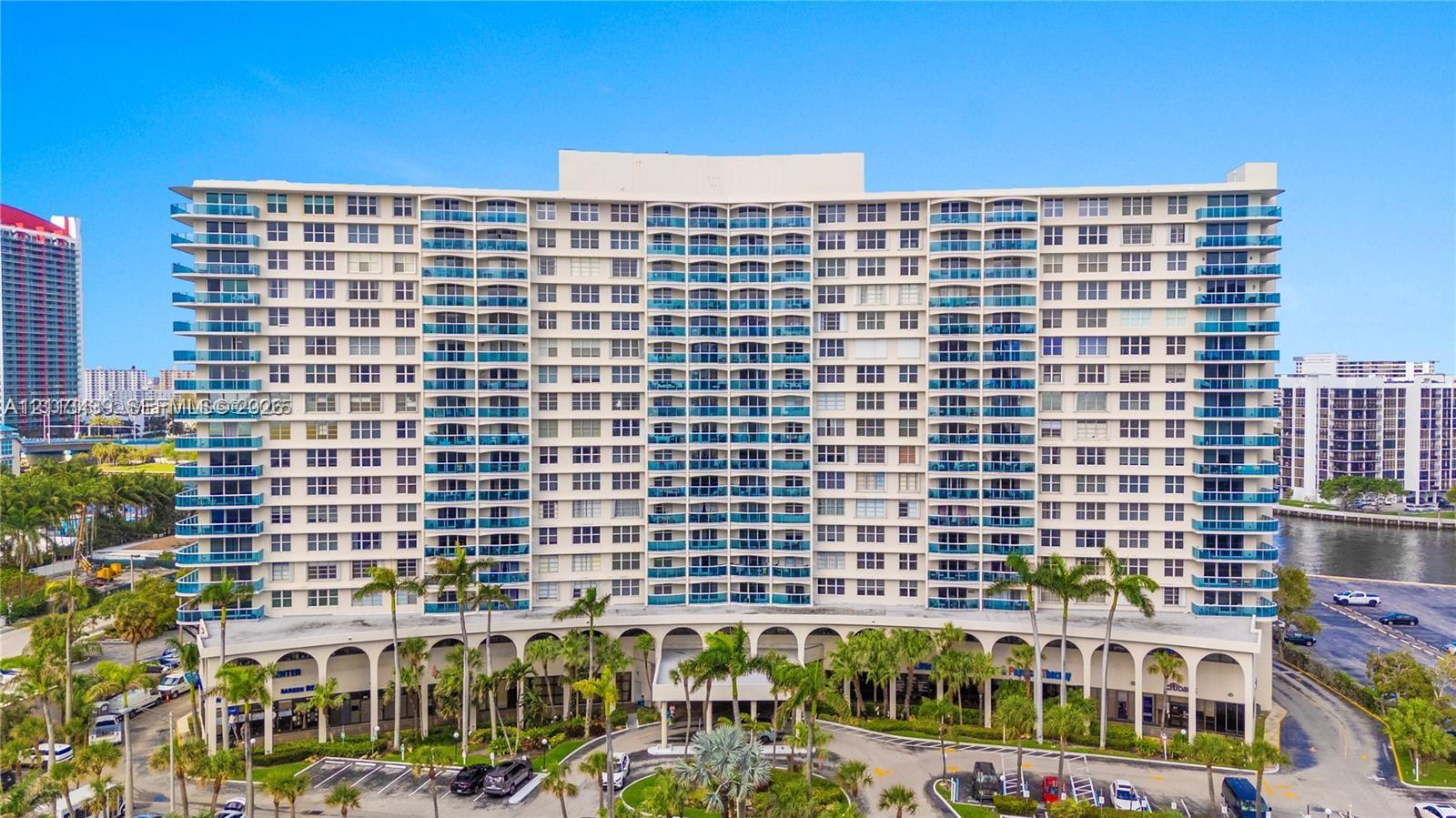 High-rise residential building featuring a curved architectural facade, private balconies with blue glass railings, an arched ground-level colonnade, and mature palm tree landscaping