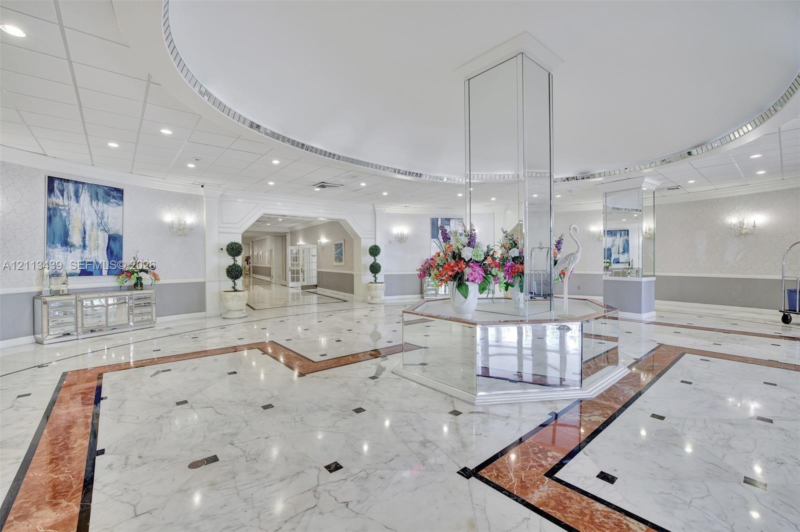 Grand foyer featuring polished stone tile flooring with decorative inlay, a central mirrored pillar, a recessed circular tray ceiling, and wall-mounted sconce lighting