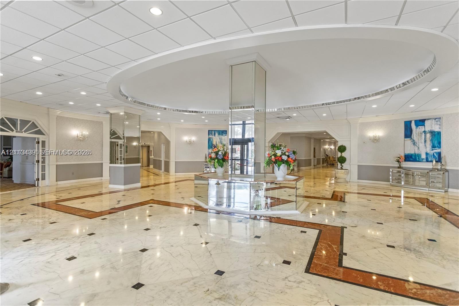 Grand lobby featuring polished stone flooring with geometric tile inlays, a central mirrored support pillar, recessed ceiling lighting, and a circular dropped ceiling detail