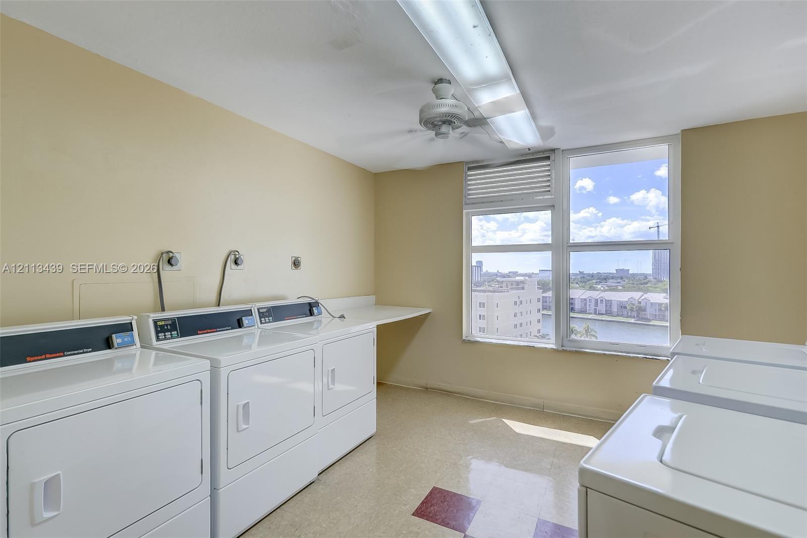 Utility room featuring multiple high-capacity washer and dryer units, expansive built-in workspace counter, large window with natural light, and ceiling-mounted lighting