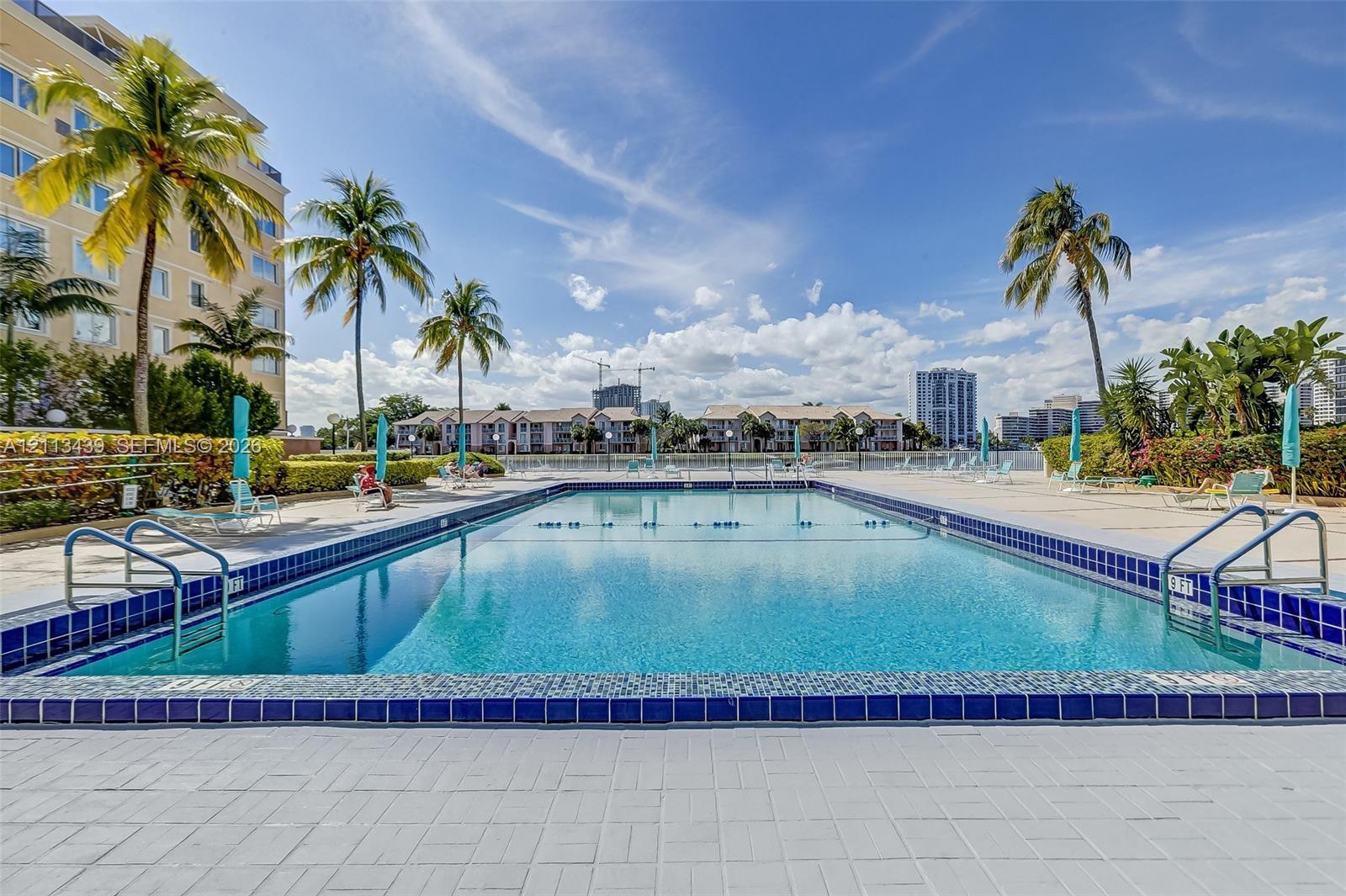 Rectangular swimming pool featuring blue ceramic tile trim, metal entry railings, and an expansive paved pool deck surrounded by palm trees and landscaped hedging